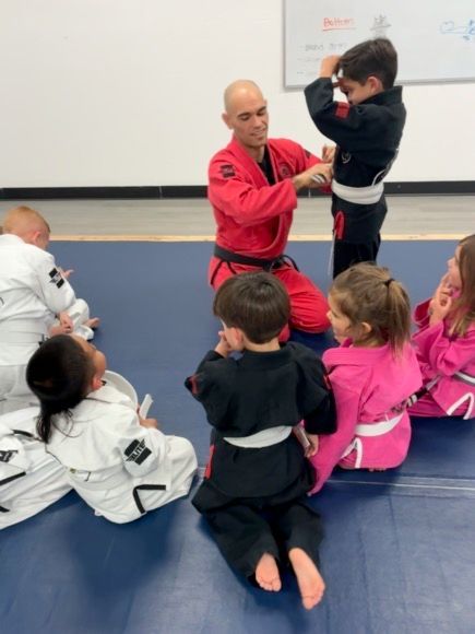 Martial arts instructor demonstrating a move to seated children in white, pink, and black uniforms