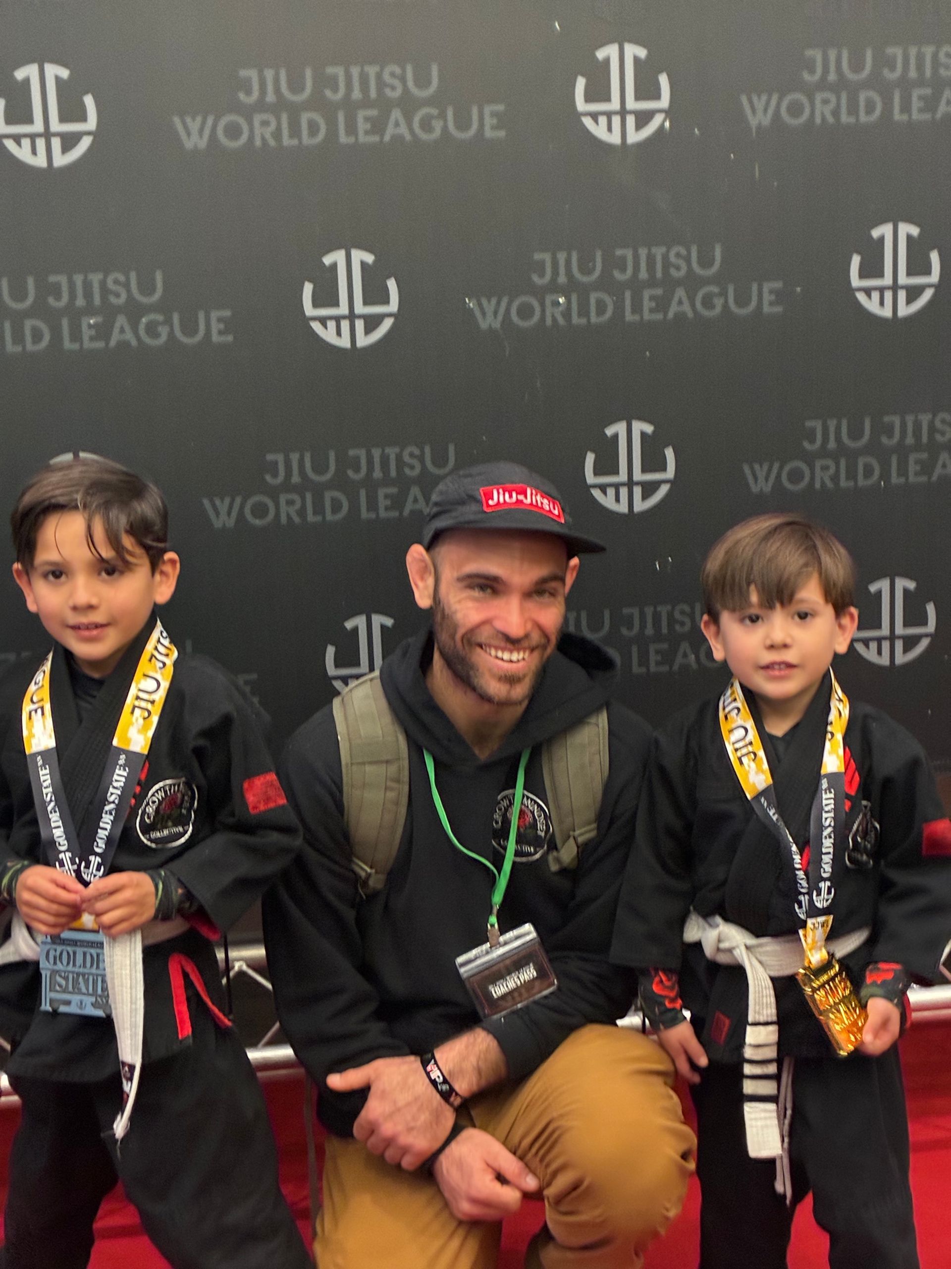 Three medalists posing with a coach at a Jiu Jitsu World League event backdrop