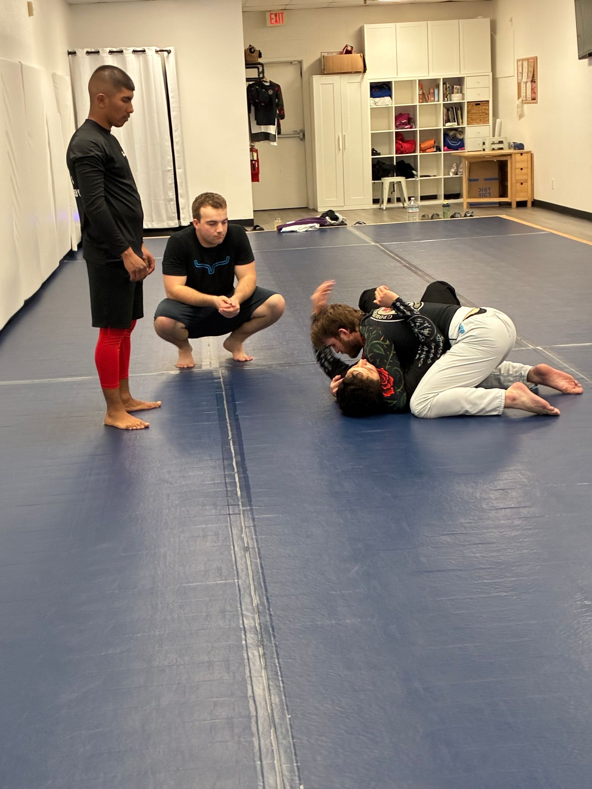 Martial arts class on blue mats, with two people grappling and two instructors watching nearby.