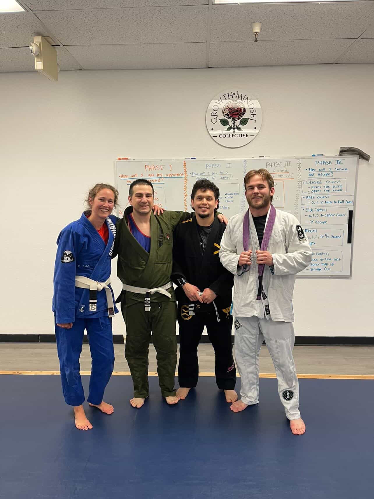 Four people in martial arts gis pose smiling on a blue mat in a dojo classroom.