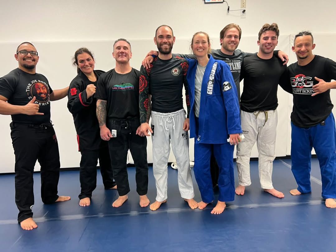 Seven smiling martial arts practitioners stand together in a gym, wearing gis and training uniforms.