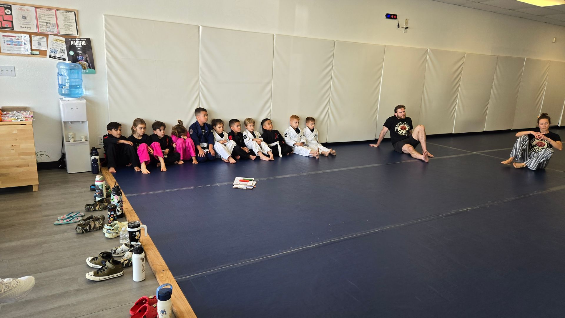 Children seated on a black mat in a martial arts studio, watching an instructor nearby