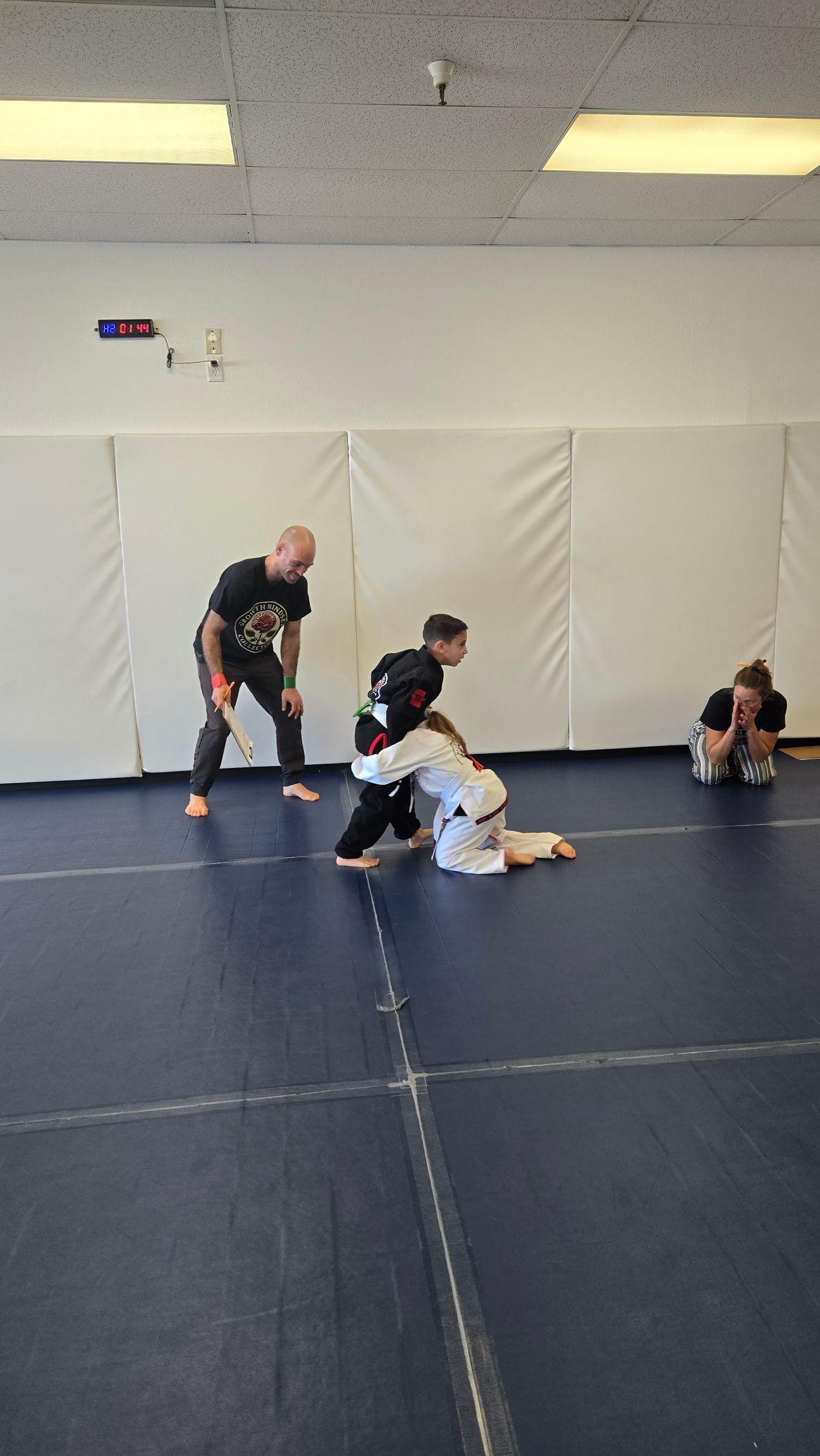 Three people practicing grappling on blue mats in a martial arts studio