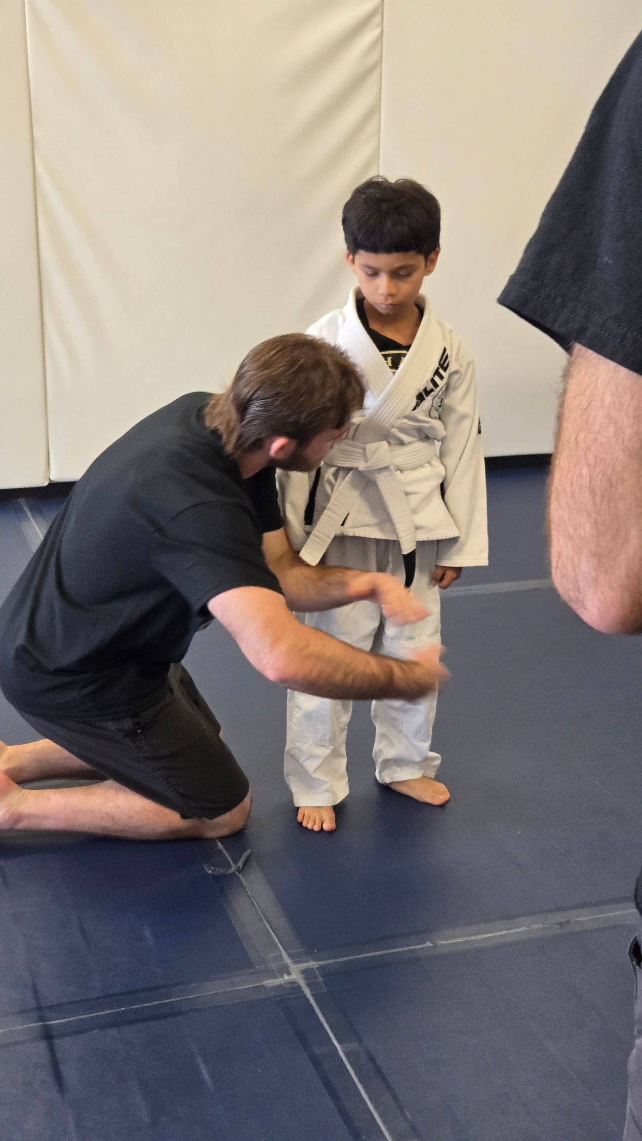 Instructor adjusts a child’s white martial arts gi on a blue mat in a dojo.