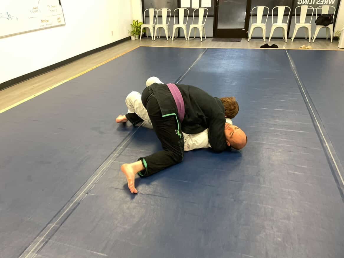 Two people grappling on a blue mat in a training room with chairs along the wall.