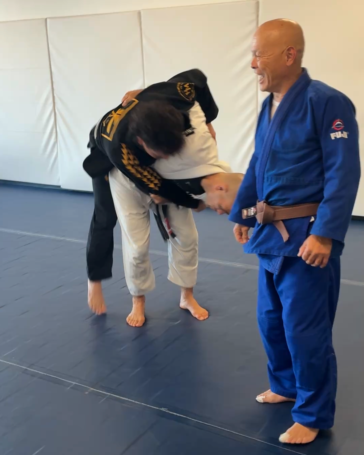 Two people practicing a judo throw on a blue mat, with one in a blue gi watching nearby