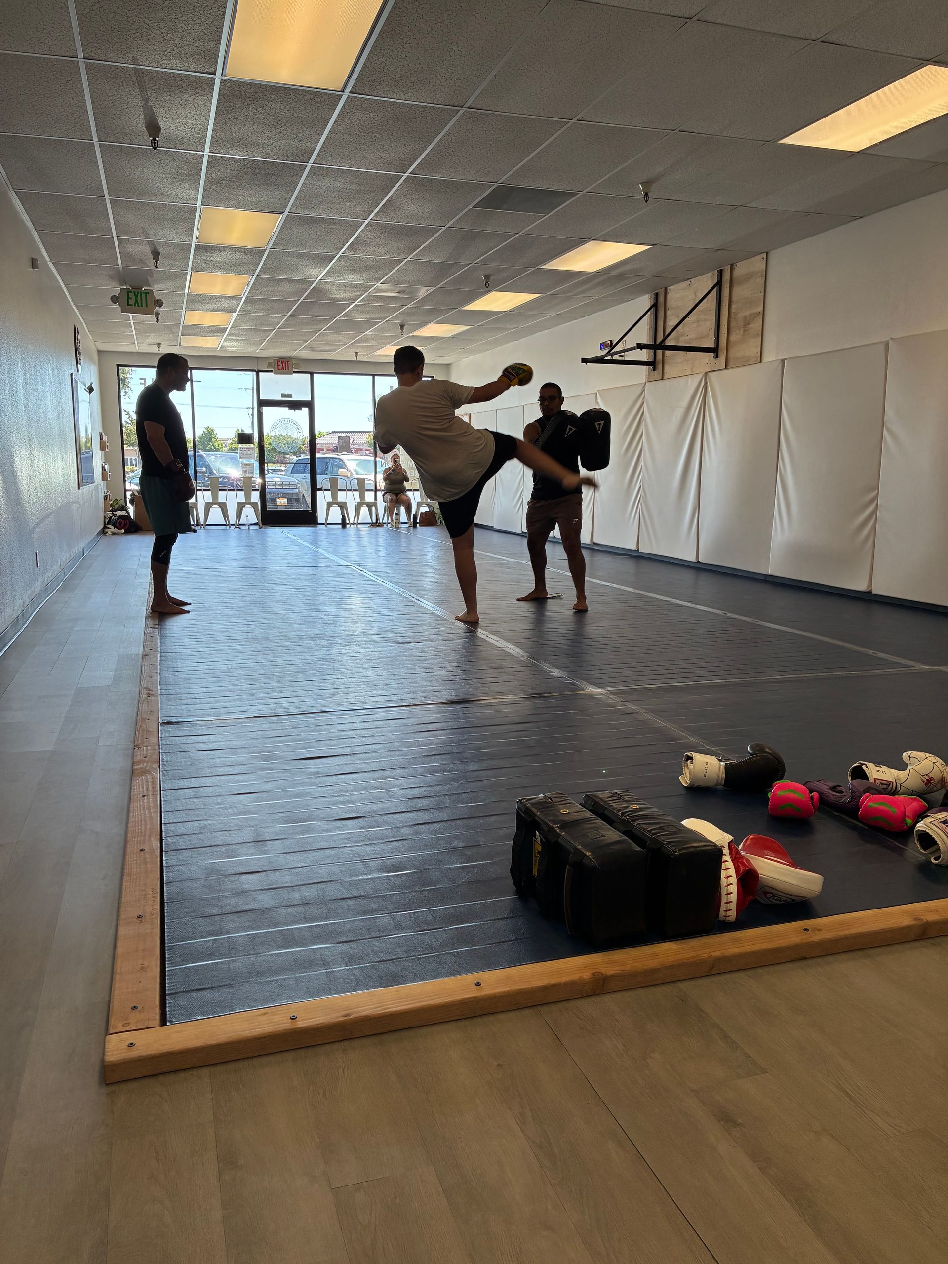 Martial arts practice in a mirrored studio with three people sparring on mats and gear on the floor.