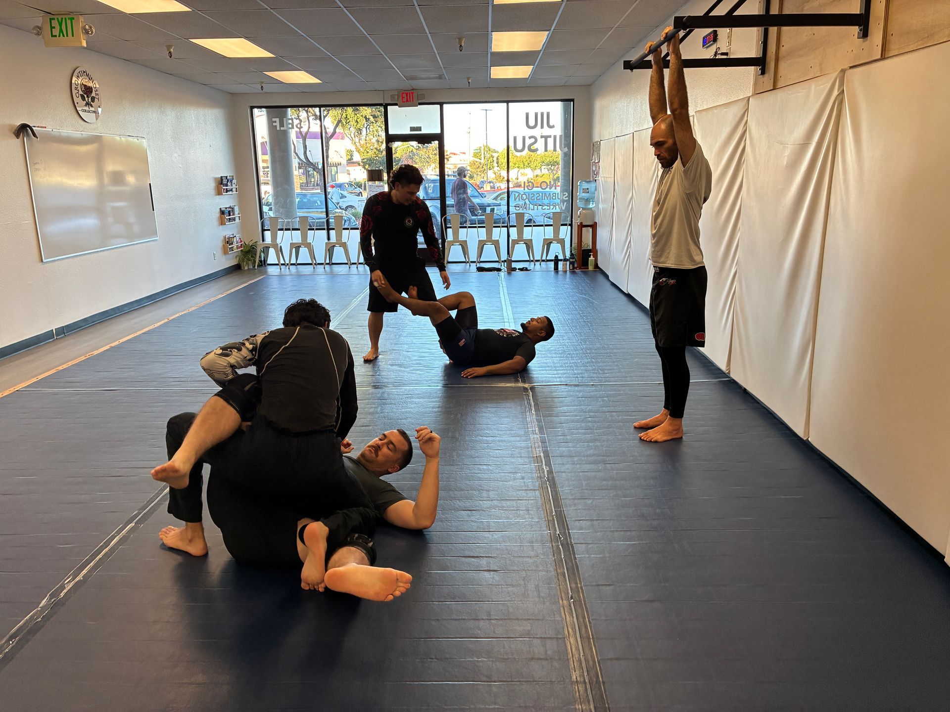 People practicing grappling on mats in a martial arts studio with mirrors and bright windows.