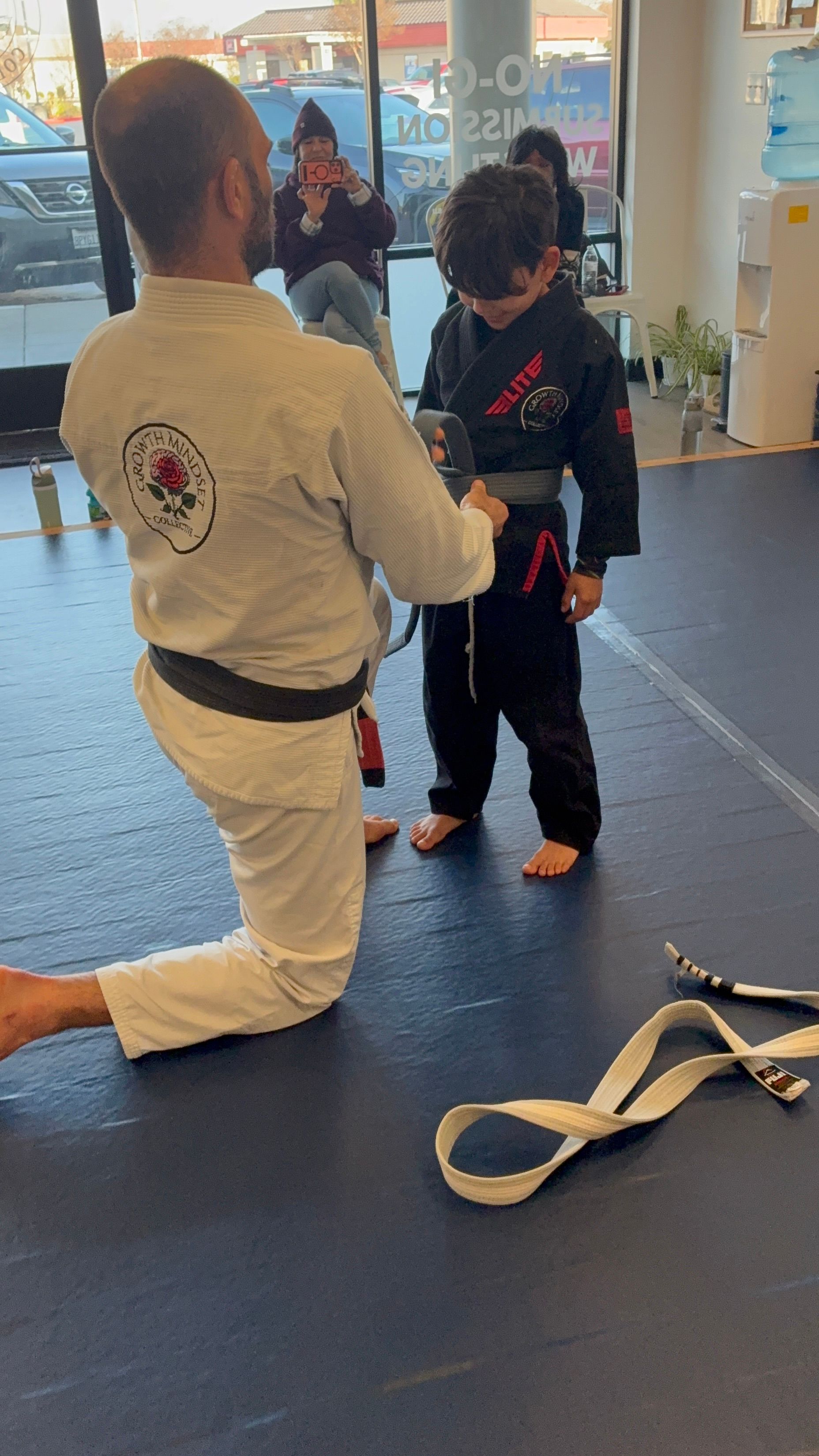 Martial arts instructor kneels beside child in a dojo, adjusting the child’s belt.