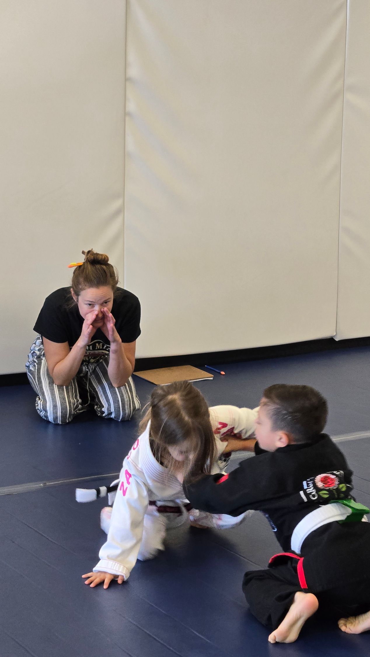 Two children on a gym mat while an adult watches nearby in a bright room