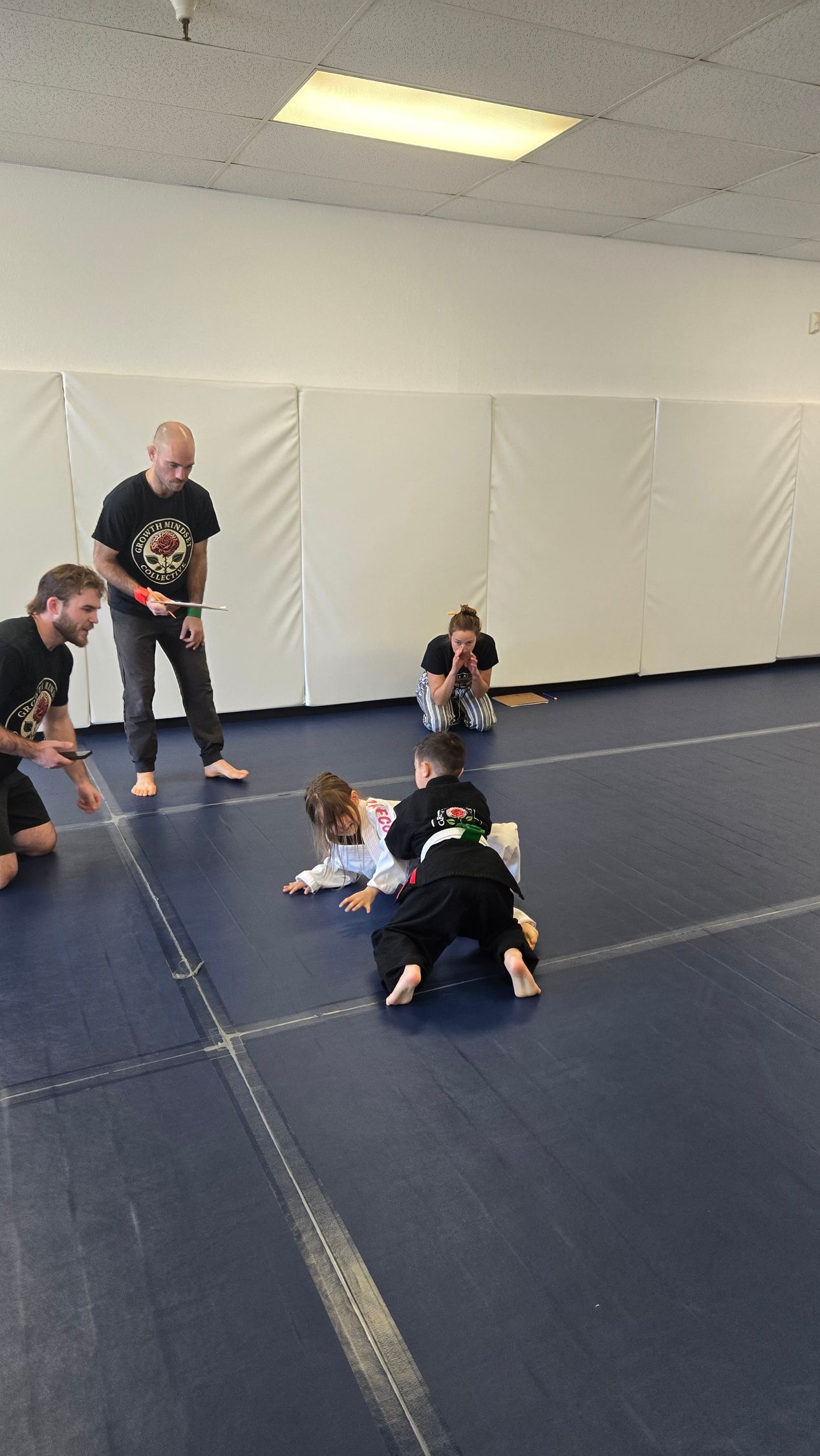 Children practicing wrestling on blue mats in a gym while a coach stands nearby.