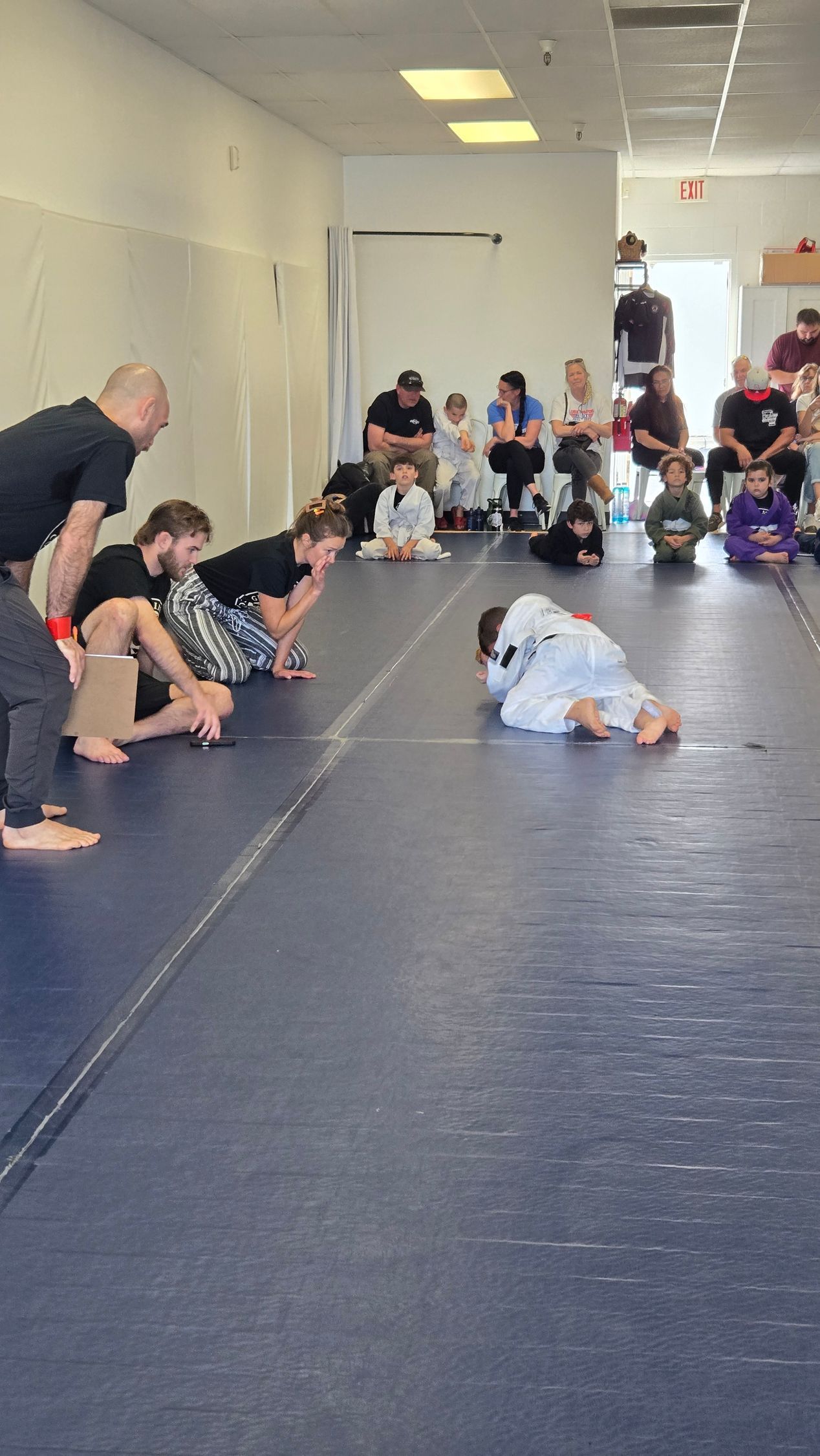 Toddler crawling on blue mat in a bright gym, with adults and children seated along the wall watching.
