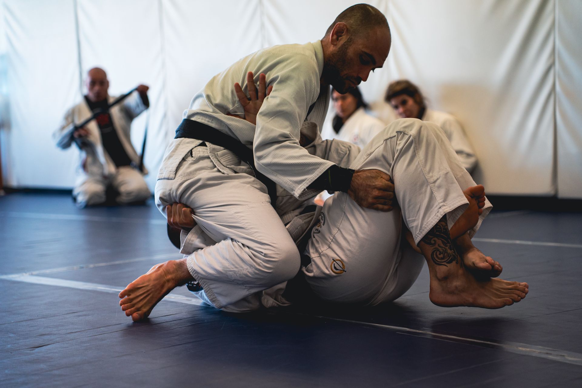 Two people in white gis practicing grappling on a blue mat in a martial arts studio