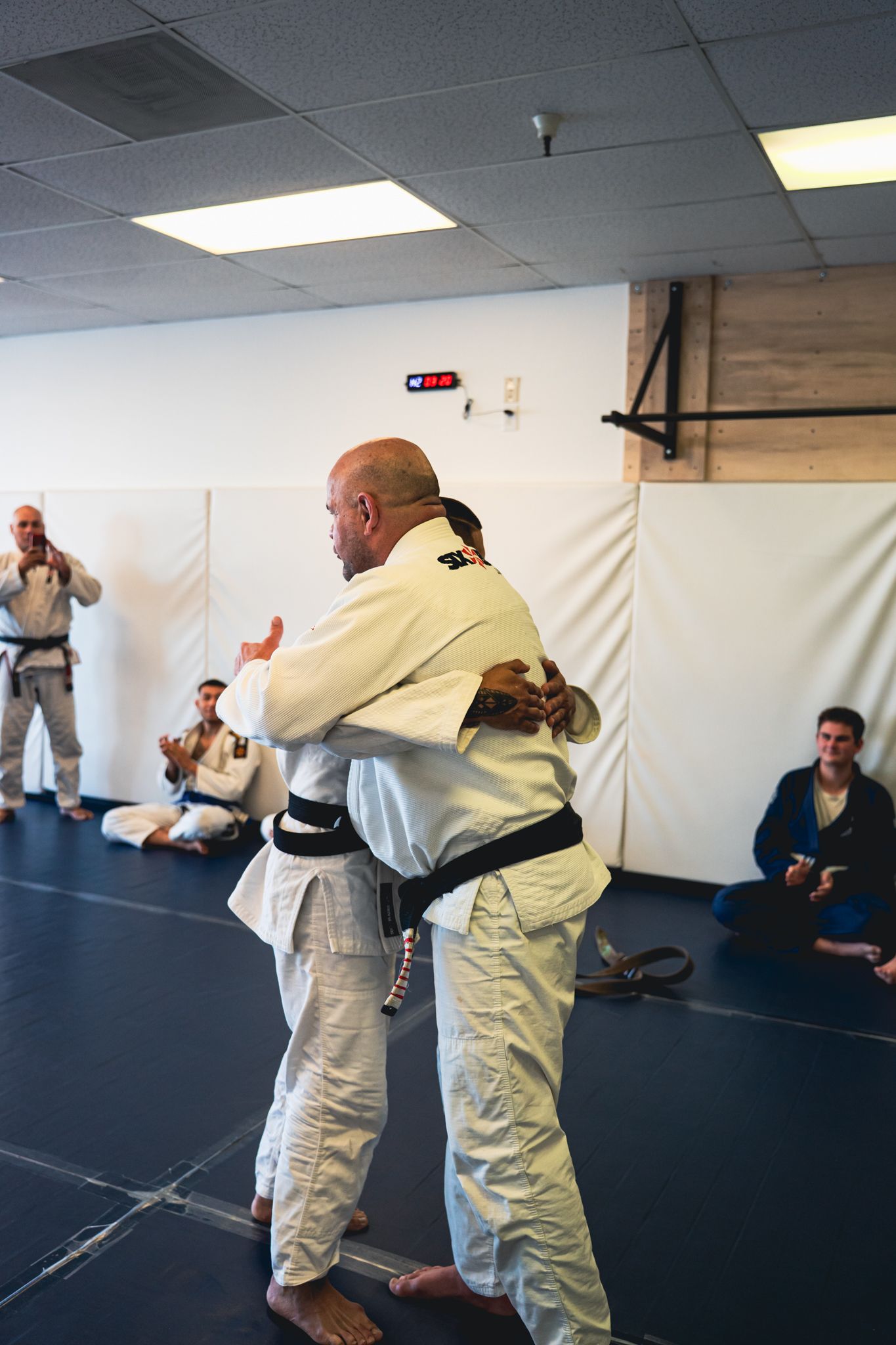 Two martial artists in white gis practicing a grappling hold in a dojo, with seated spectators watching.