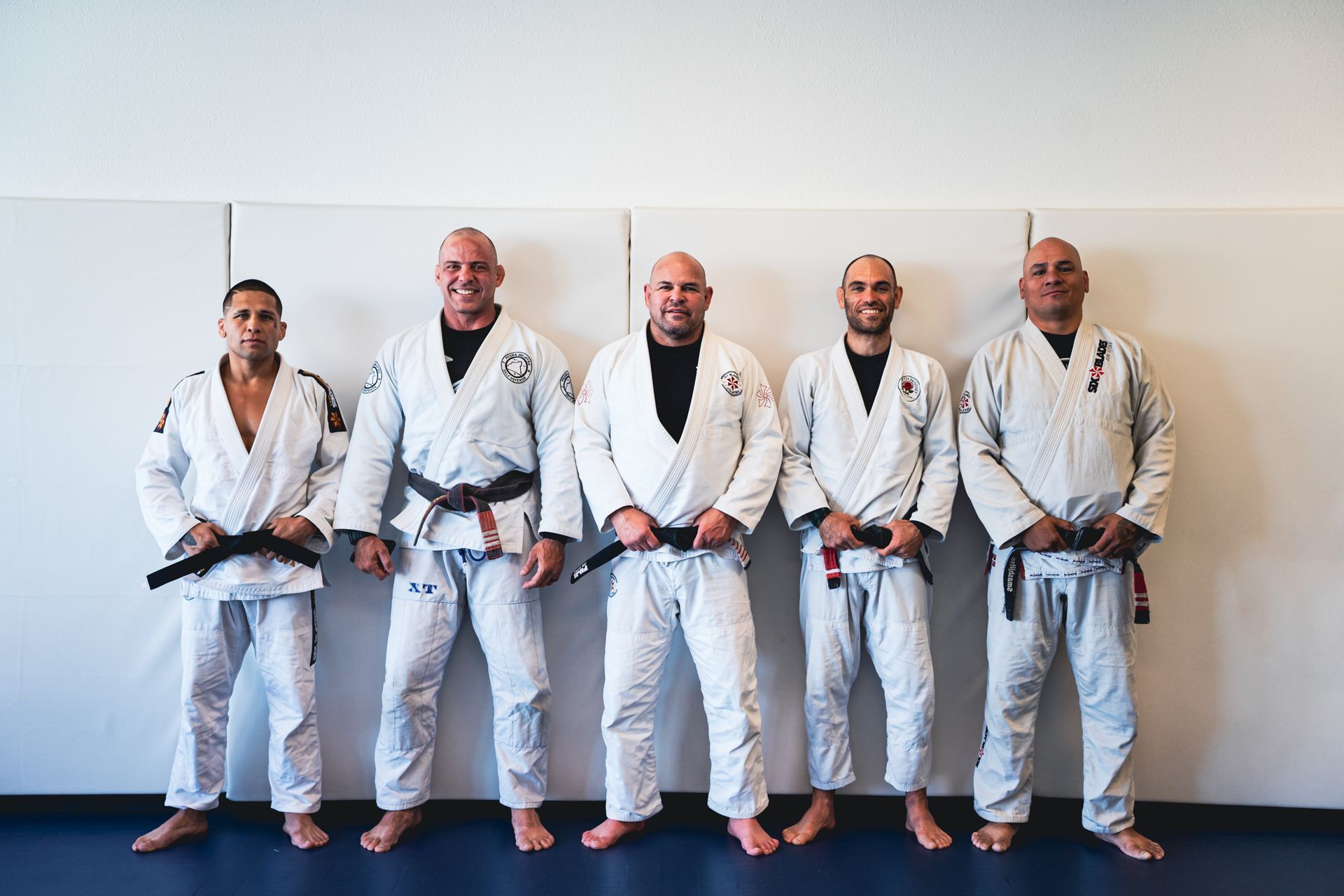 Five people in white martial arts uniforms standing side by side against a wall