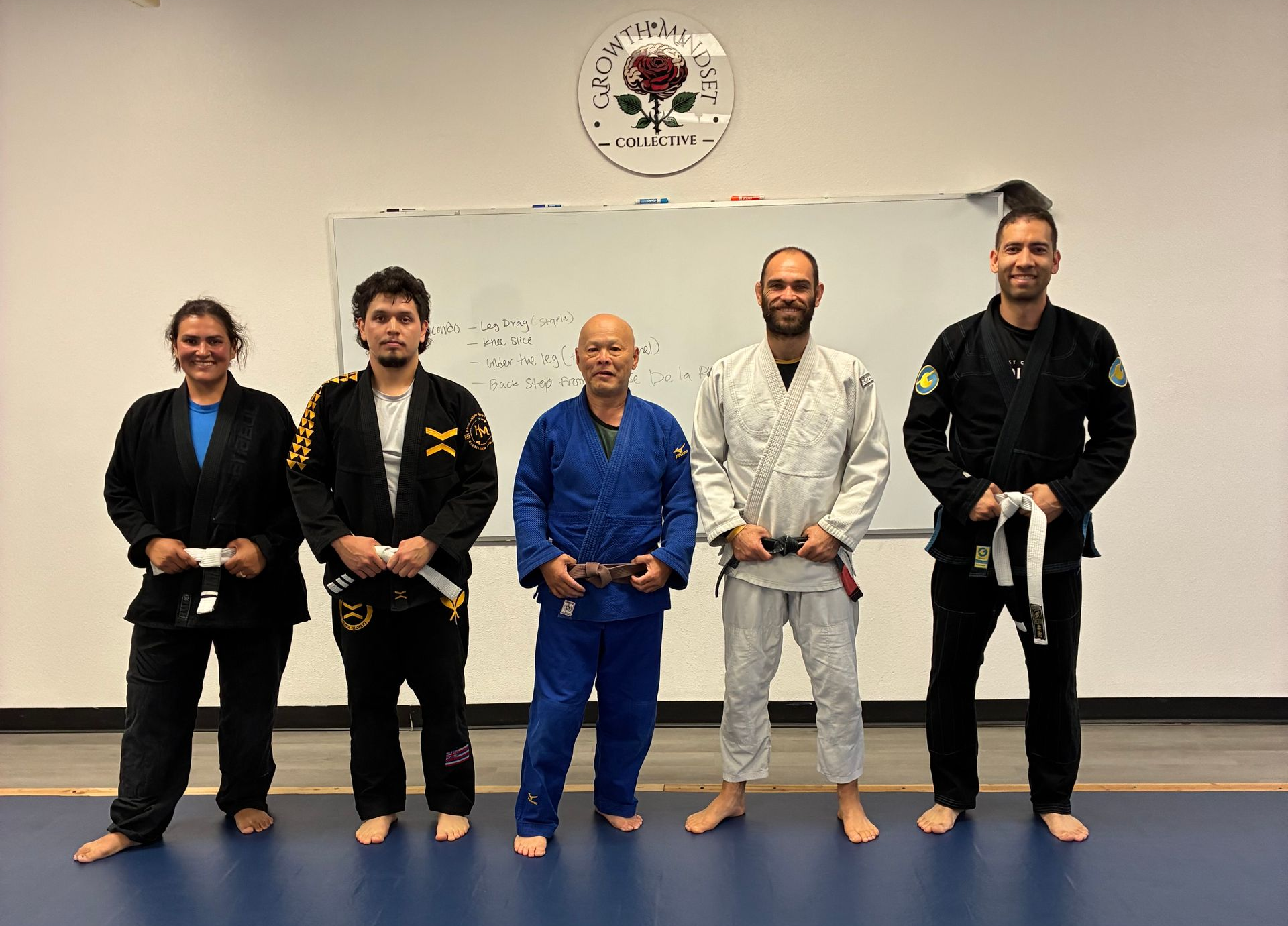 Five martial arts practitioners in gi uniforms stand barefoot in a studio holding belts after training.