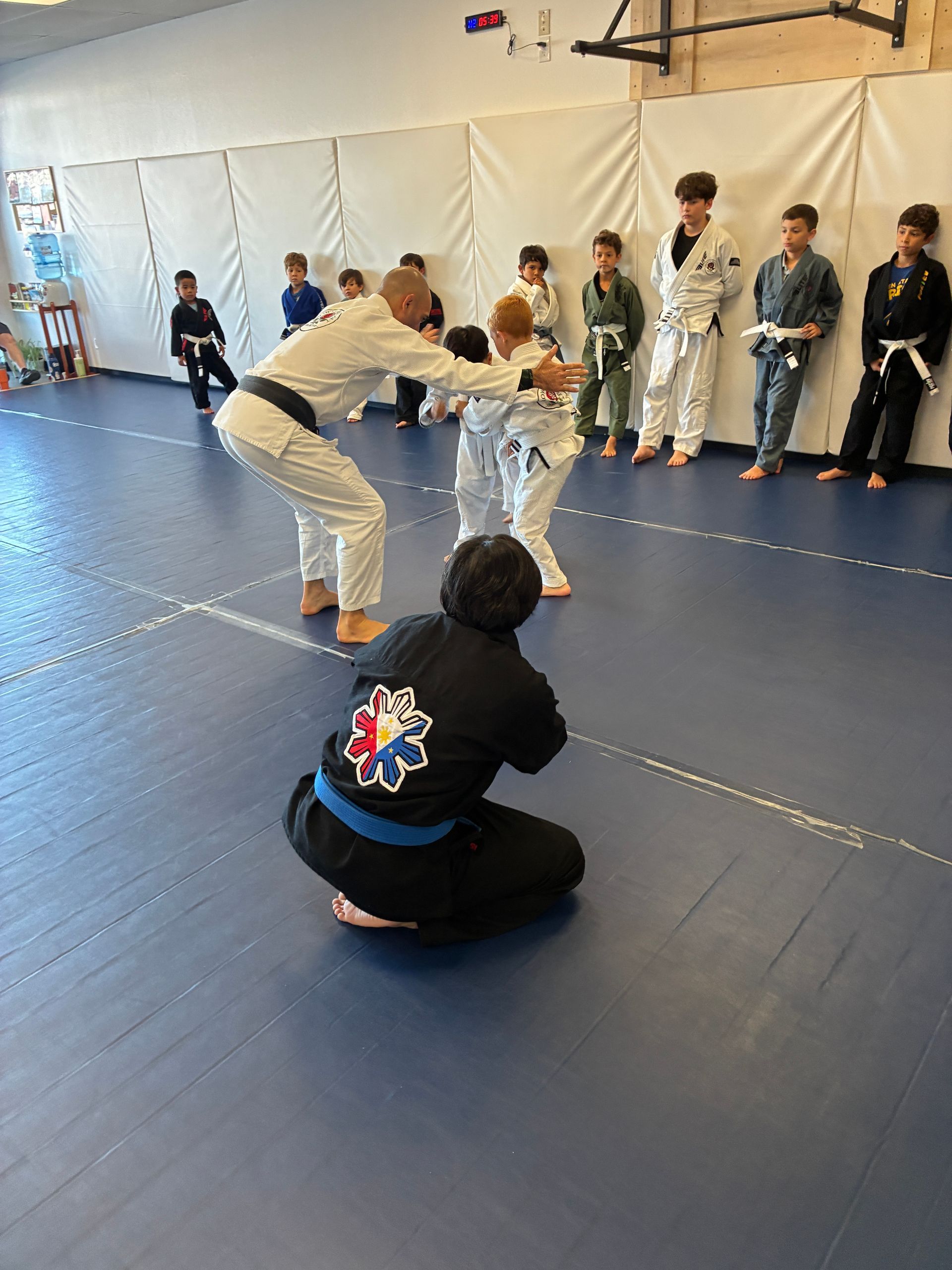 Two martial artists spar on blue mats while classmates watch along a white wall