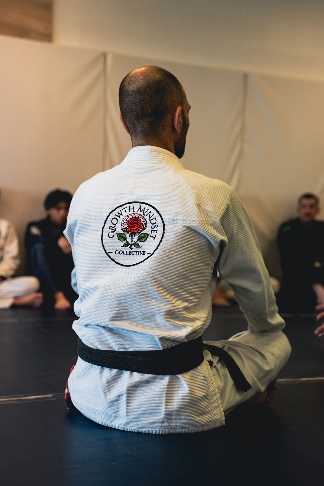 Person in a white martial arts gi sits on a mat facing a seated group in a training room.