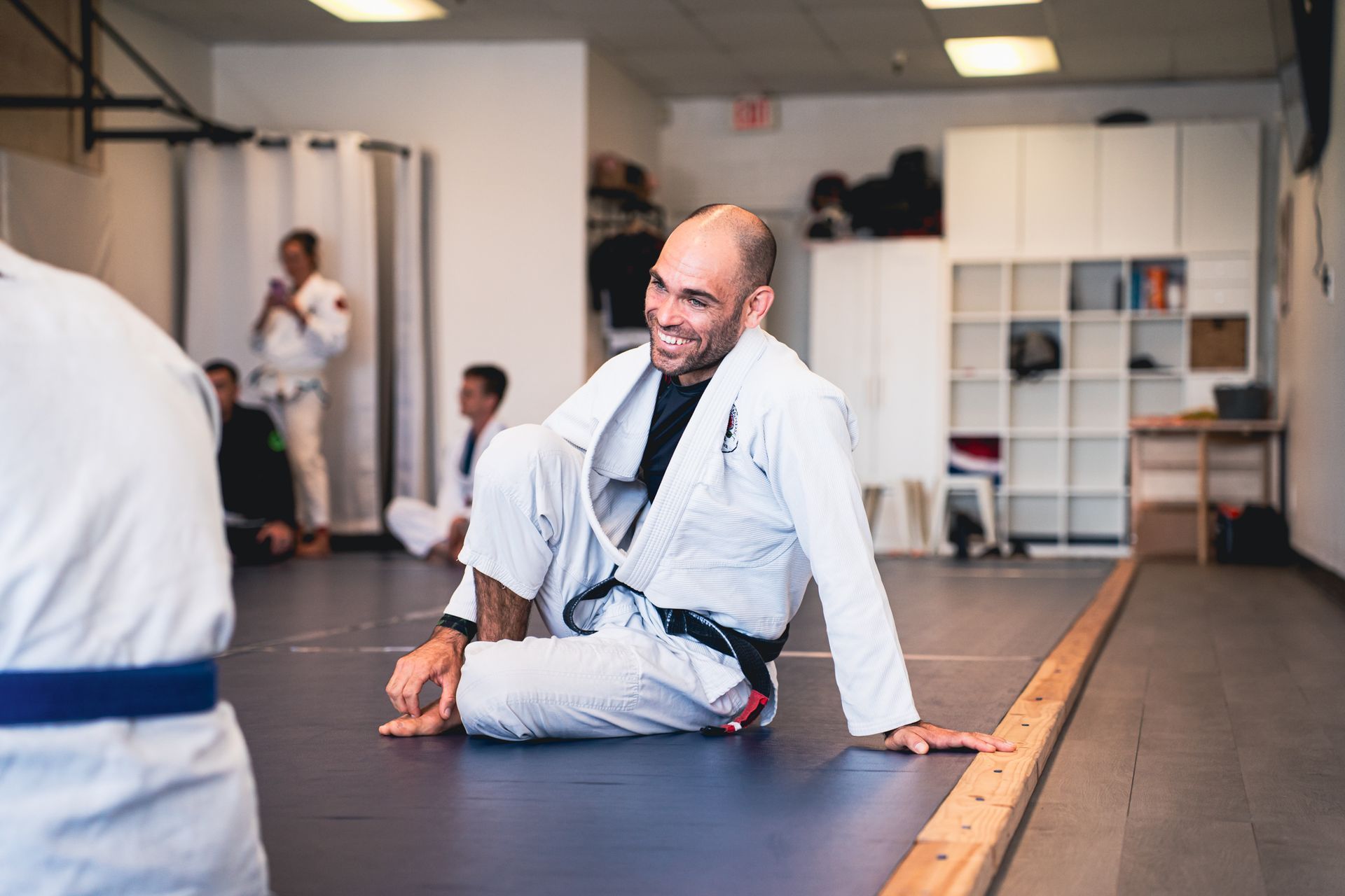 Man seated on a martial arts mat in a dojo, smiling and resting beside others in the background