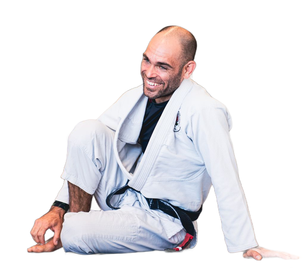 Man in a white martial arts gi sitting on the floor, smiling in a relaxed pose.