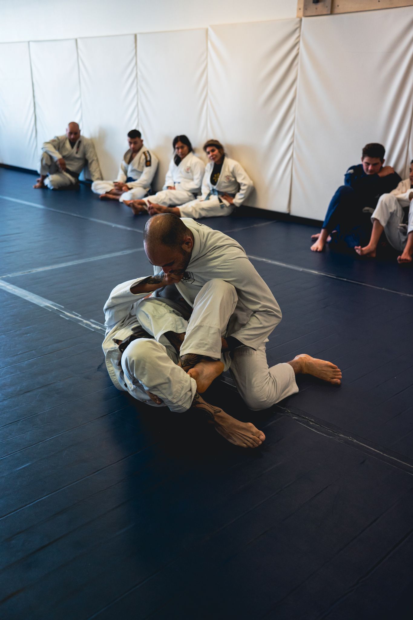 Two people practicing grappling on mats while others sit against a white wall in a martial arts studio