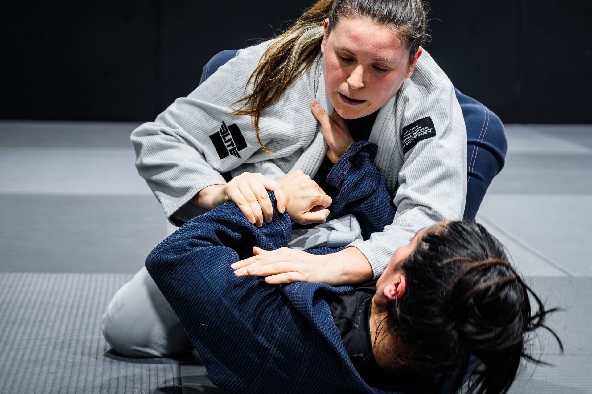 Two women in grappling uniforms practicing a ground hold on a mat, with one pinning the other.