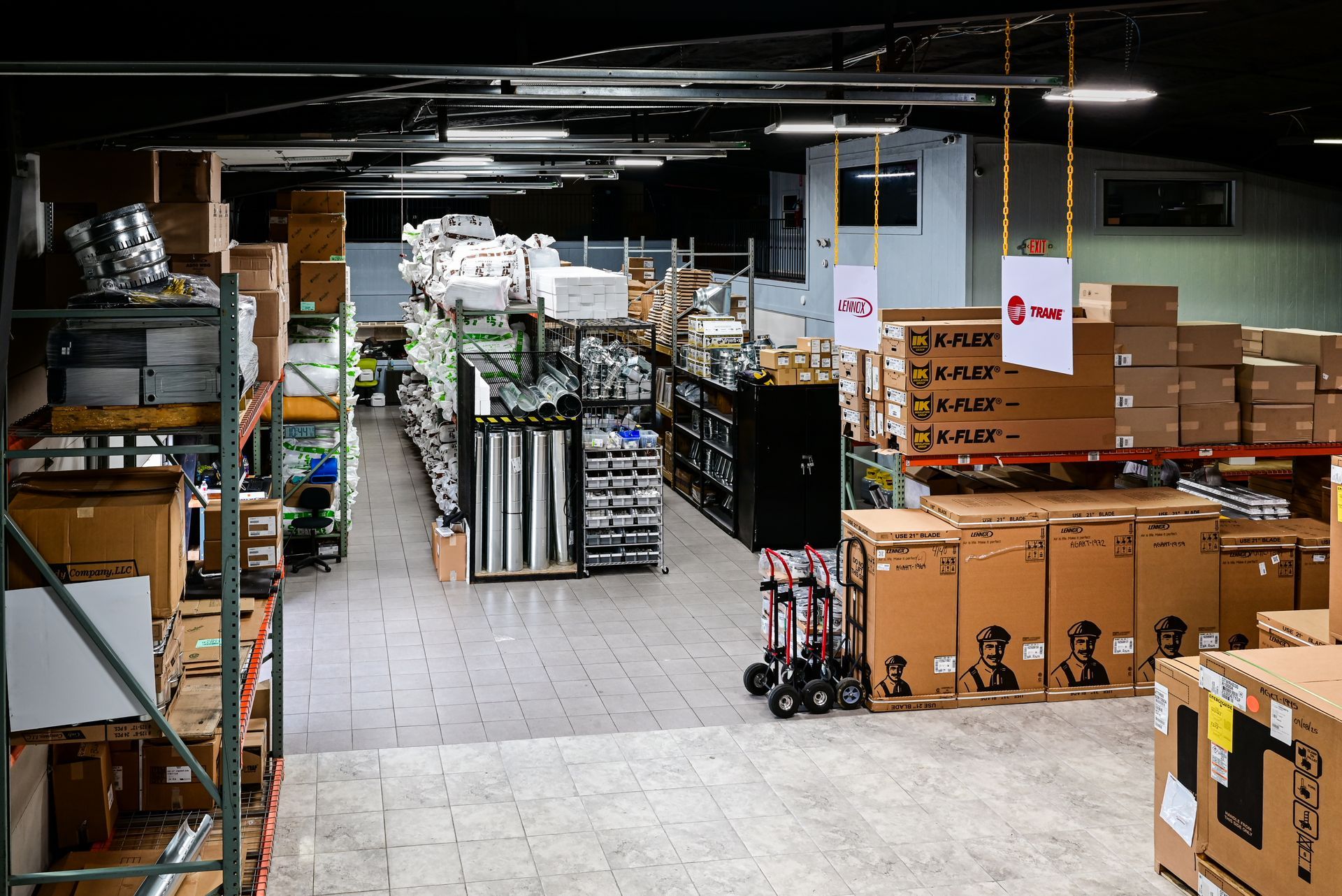A cluttered warehouse aisle with stacked cardboard boxes, shelves, and pallets under fluorescent lighting