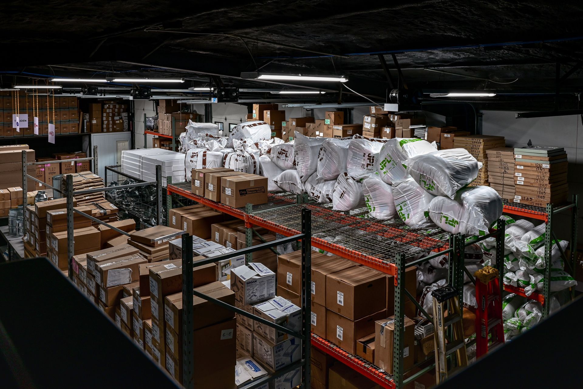 Warehouse filled with stacked boxes, wrapped rolls, and pallets under dim industrial lighting