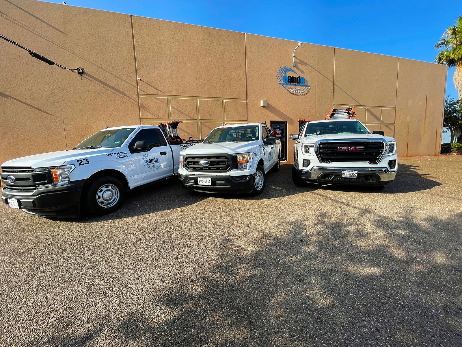 Three white service trucks parked on gravel in front of a tan building under a clear blue sky