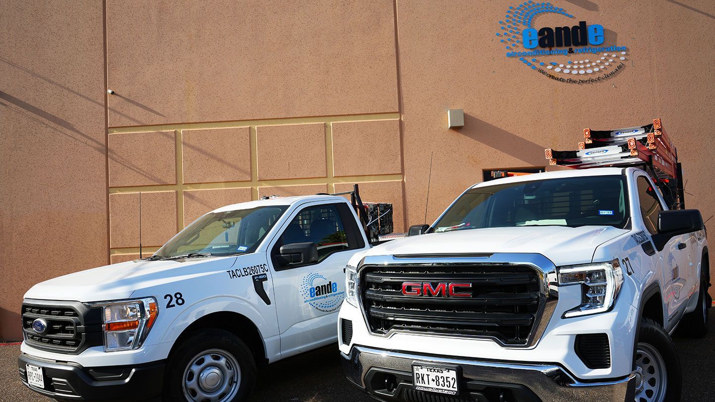 Two white service trucks parked in front of a tan building, one with a lift on top.