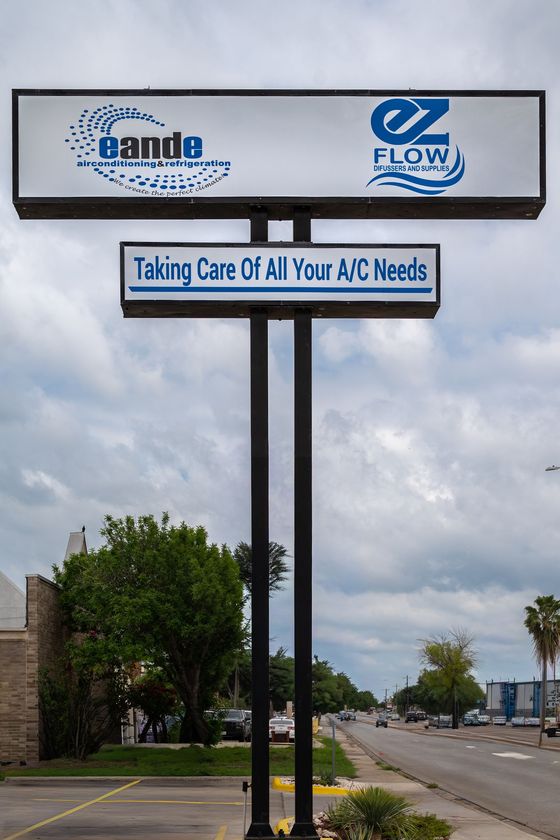 A roadside AC service sign with blue logos and the slogan “Taking Care of All Your AC Needs” under cloudy skies