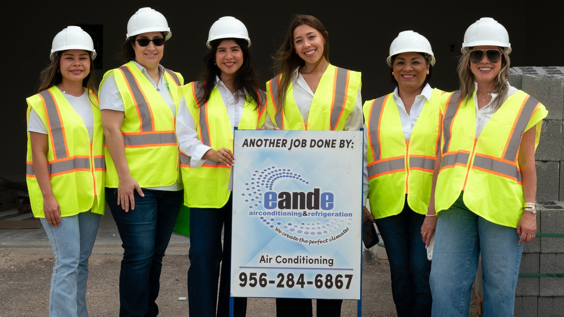 Group of six workers in white hard hats and yellow safety vests standing behind a sign outdoors.