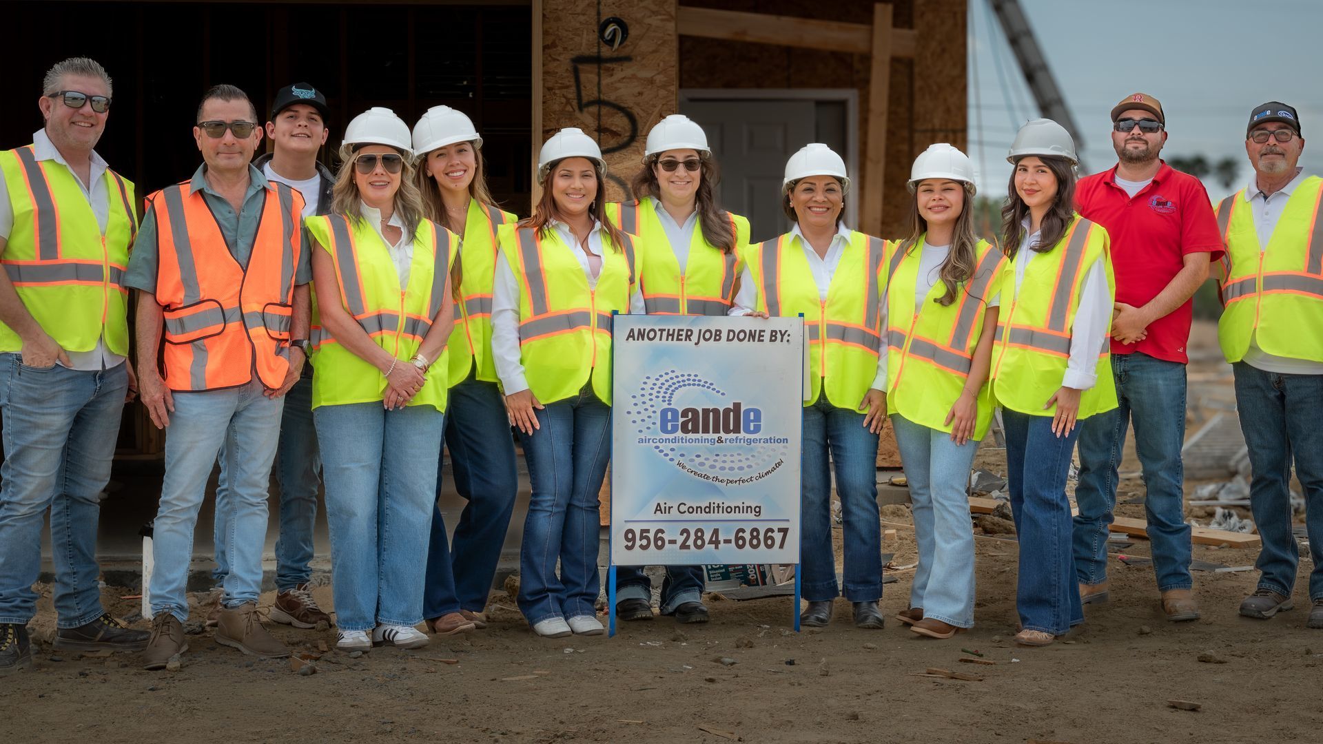 Construction crew in hard hats and safety vests posing at a building site with a sign.