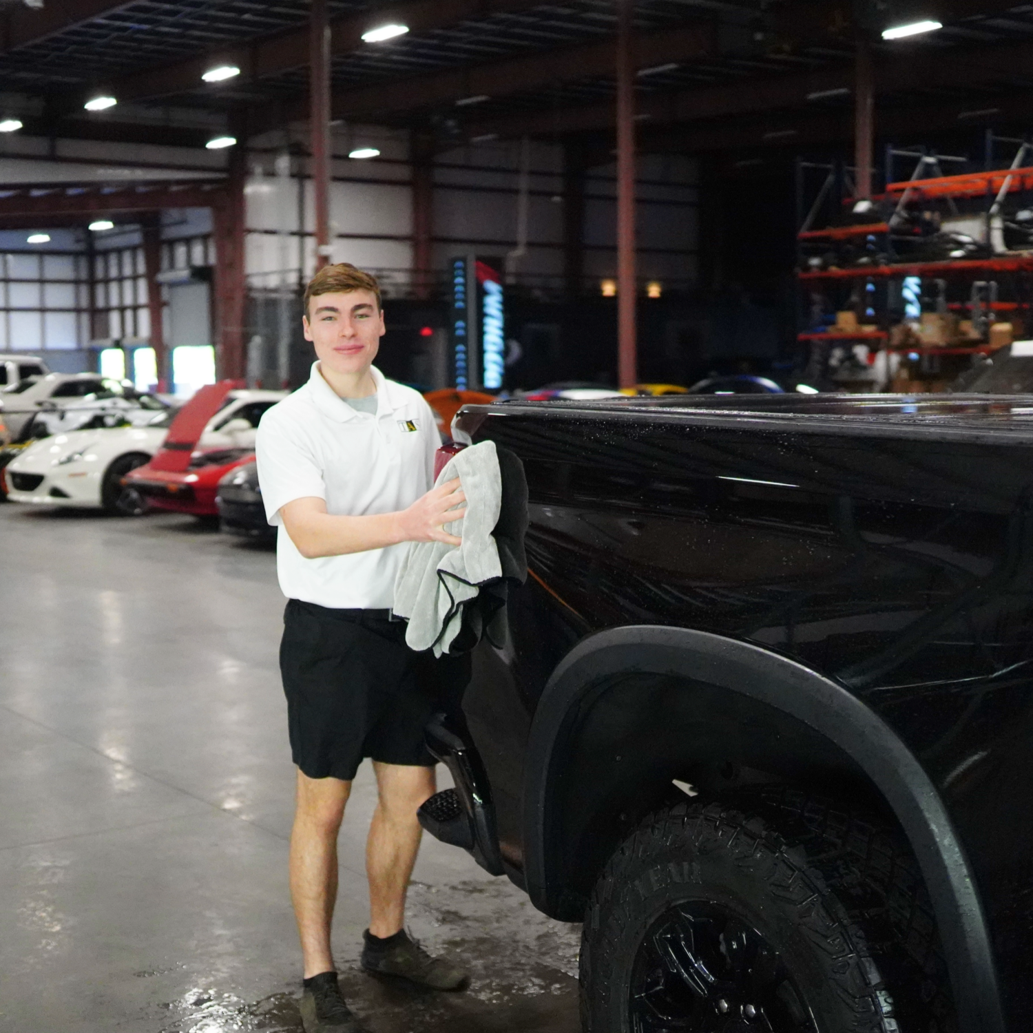 Man washing a black truck in a car warehouse, wearing a white shirt and black shorts.