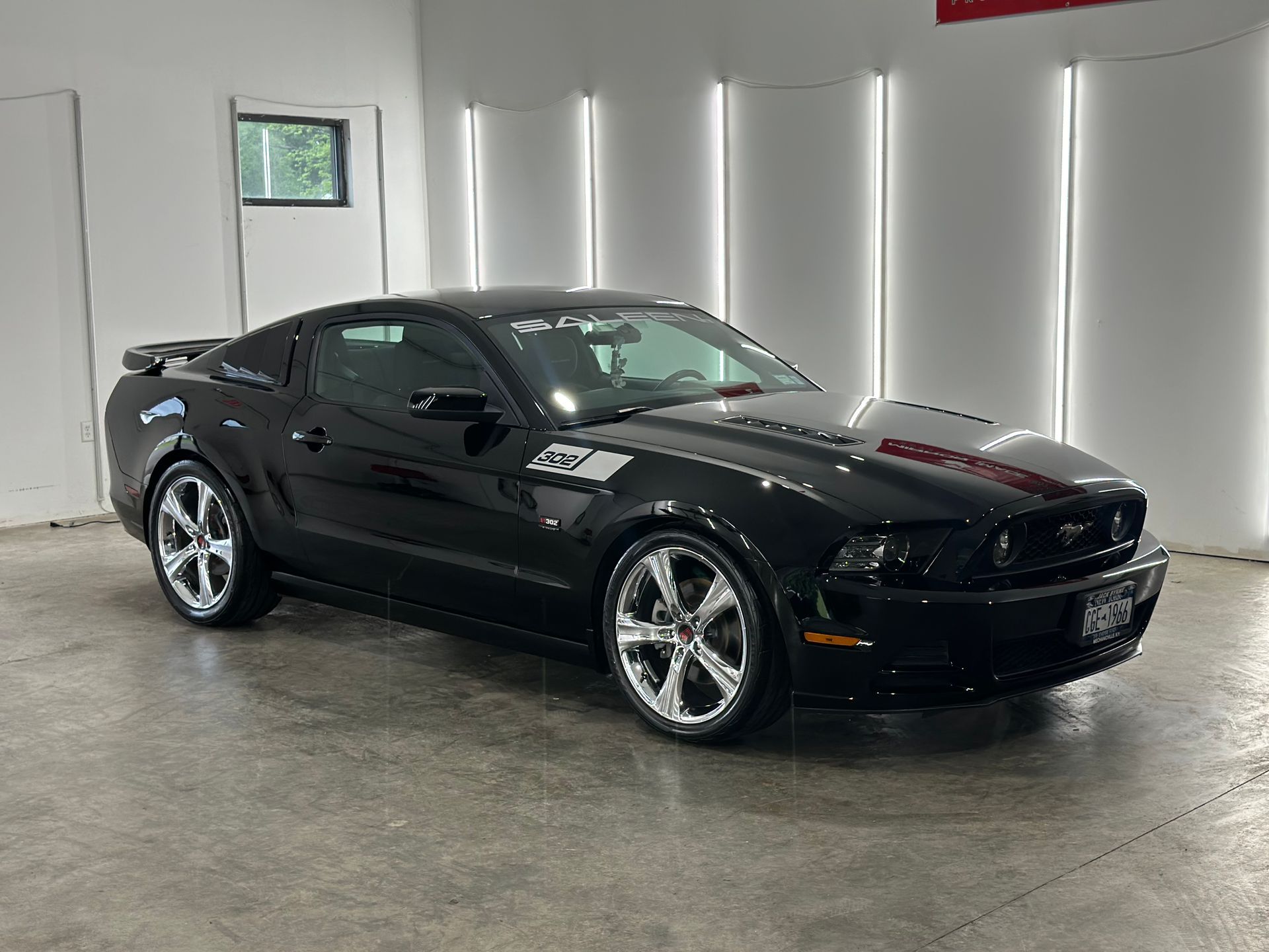 A black Ford Mustang parked in a brightly lit professional garage or showroom with white walls and vertical lighting.
