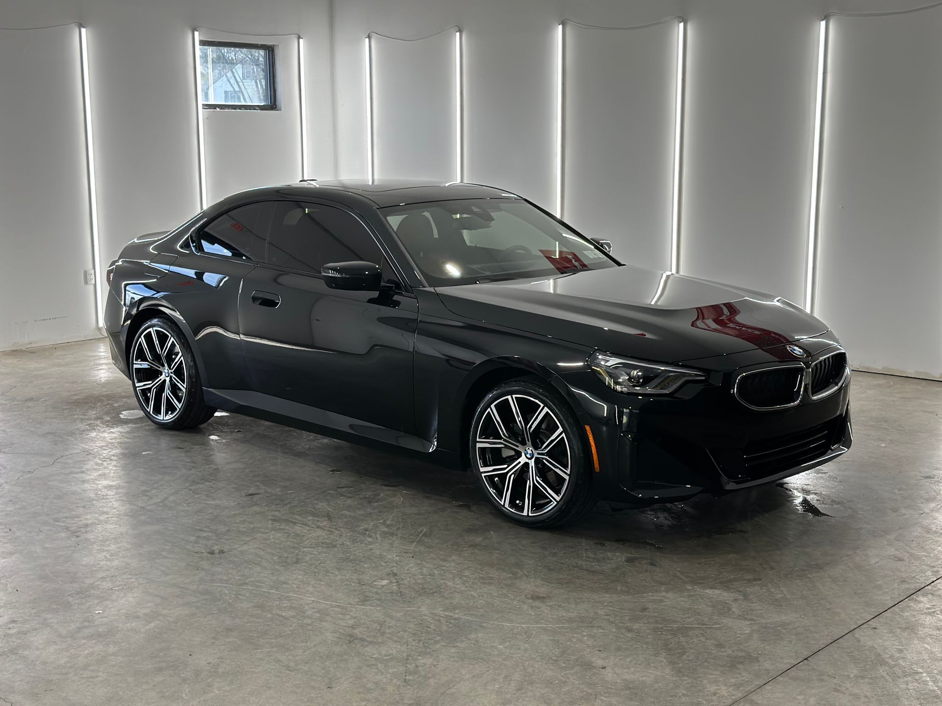 A sleek black BMW 2 Series coupe parked in a bright, minimalist studio with vertical LED lighting.