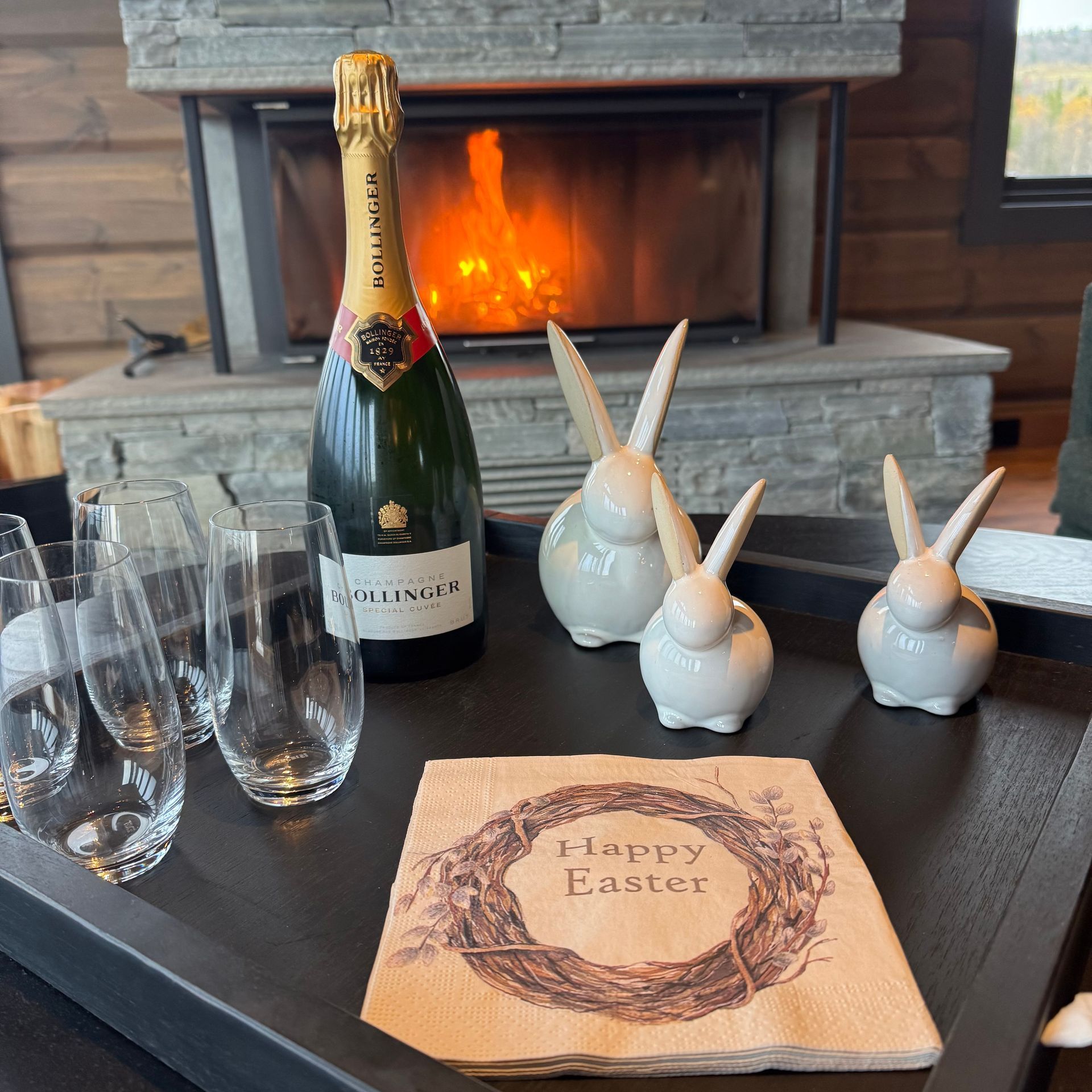 Champagne bottle, glasses, Easter bunnies, and napkins on a tray near a fireplace.