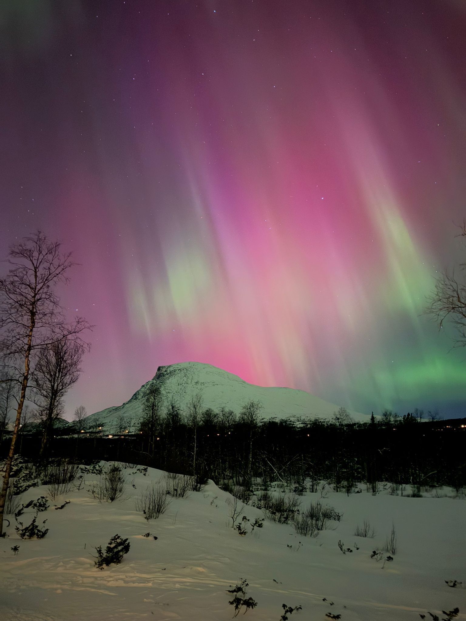 Aurora borealis lights up the night sky with pink and green over a snowy landscape and mountain.