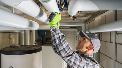Plumber in a basement, wearing gloves, working on overhead pipes near a water heater.