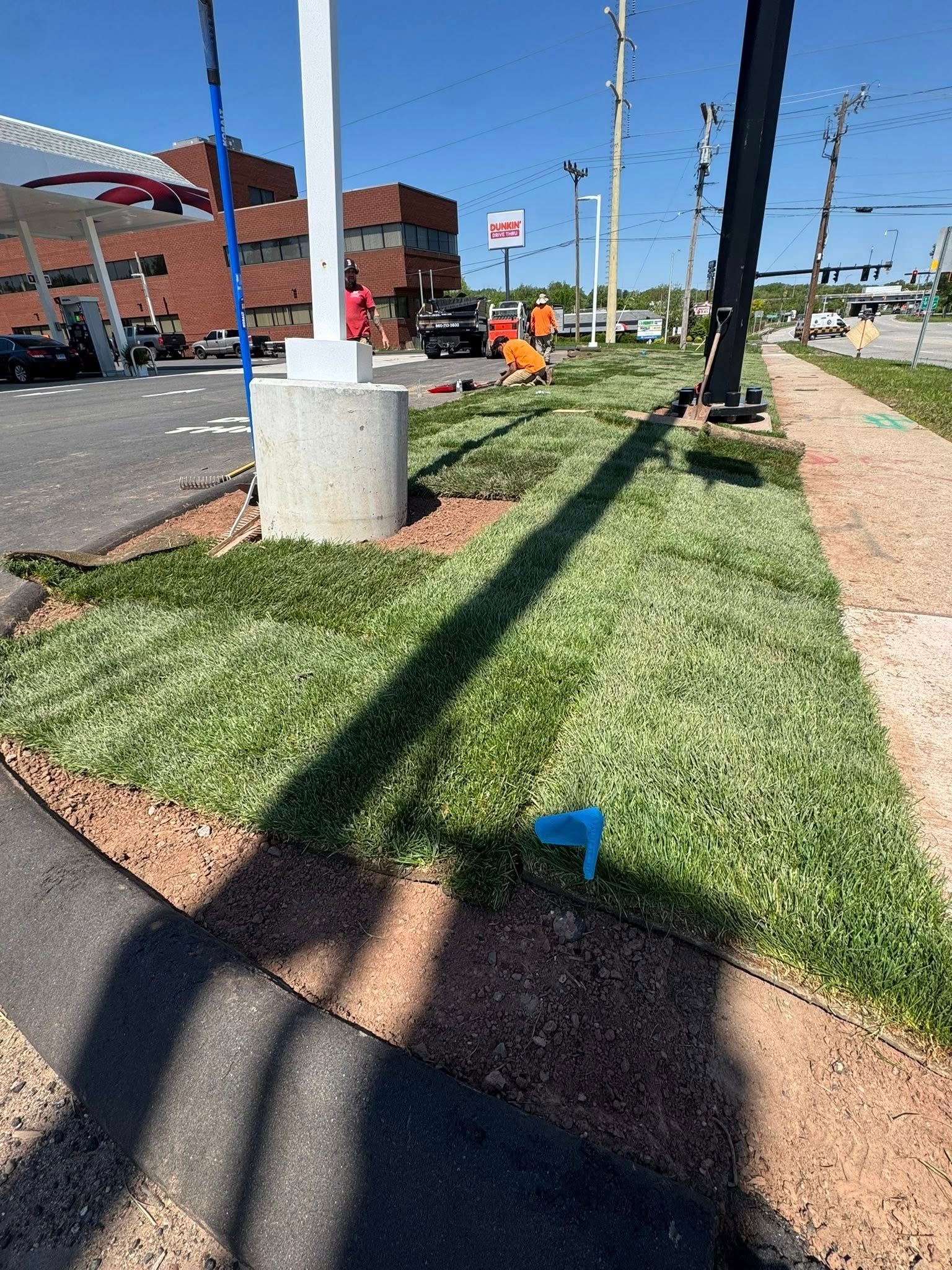 Sidewalk with a grassy strip, concrete light pole, workers in orange vests, and blue utility flags along the street.