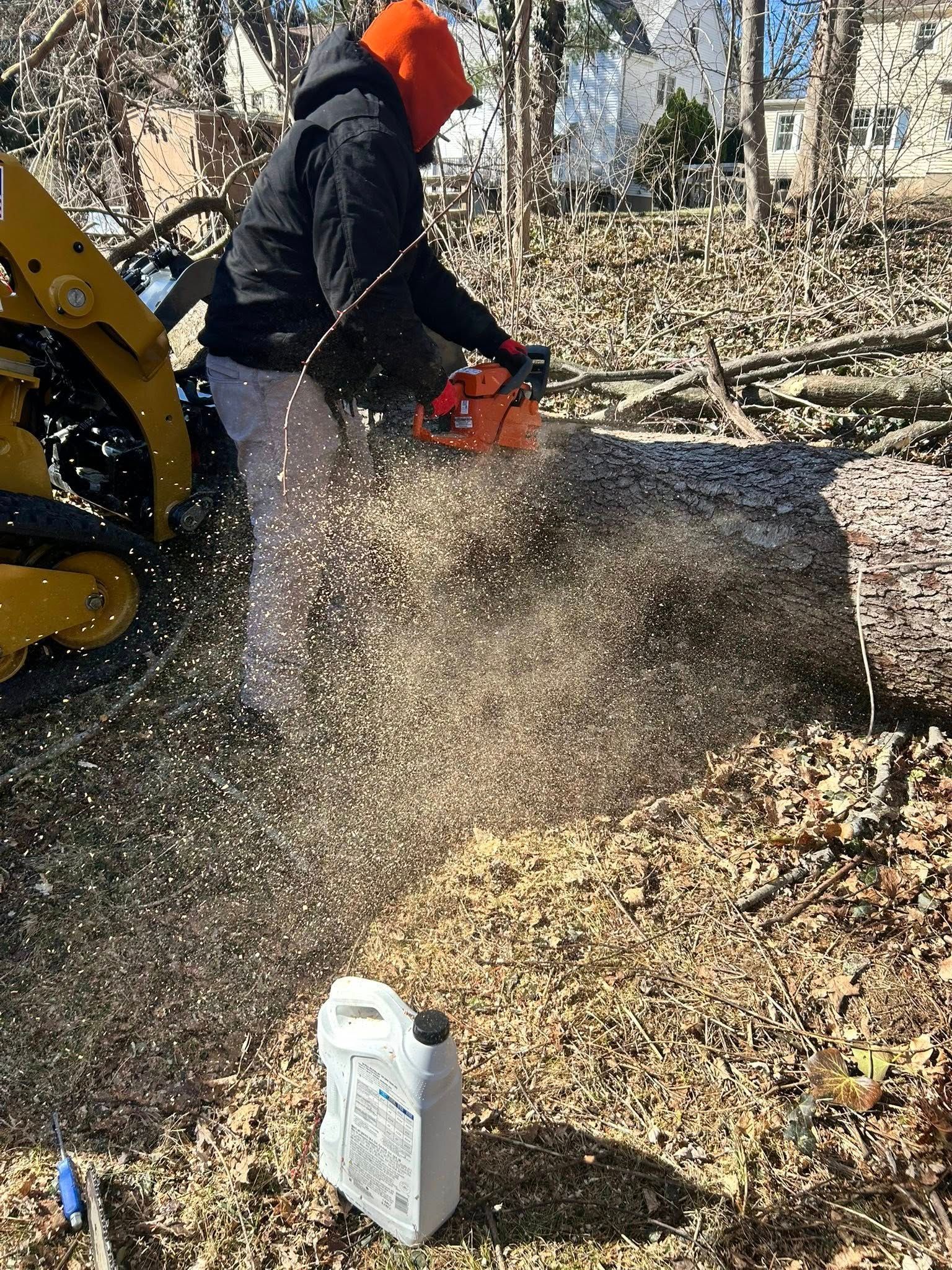 Person in orange hood using a chainsaw to cut a log outdoors, with wood chips flying.