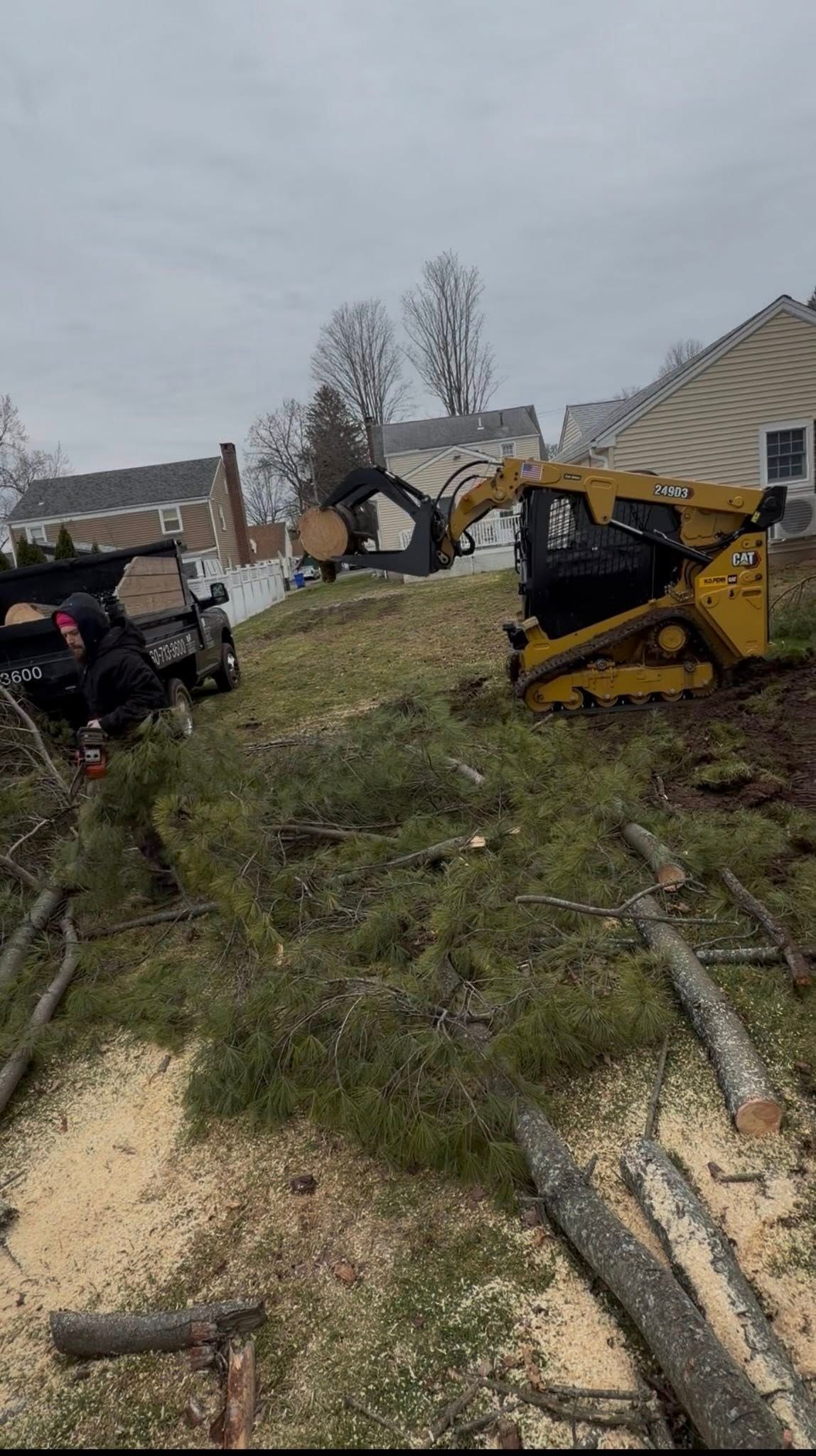 Yellow skid steer on muddy grassy slope near houses under gray sky after a storm