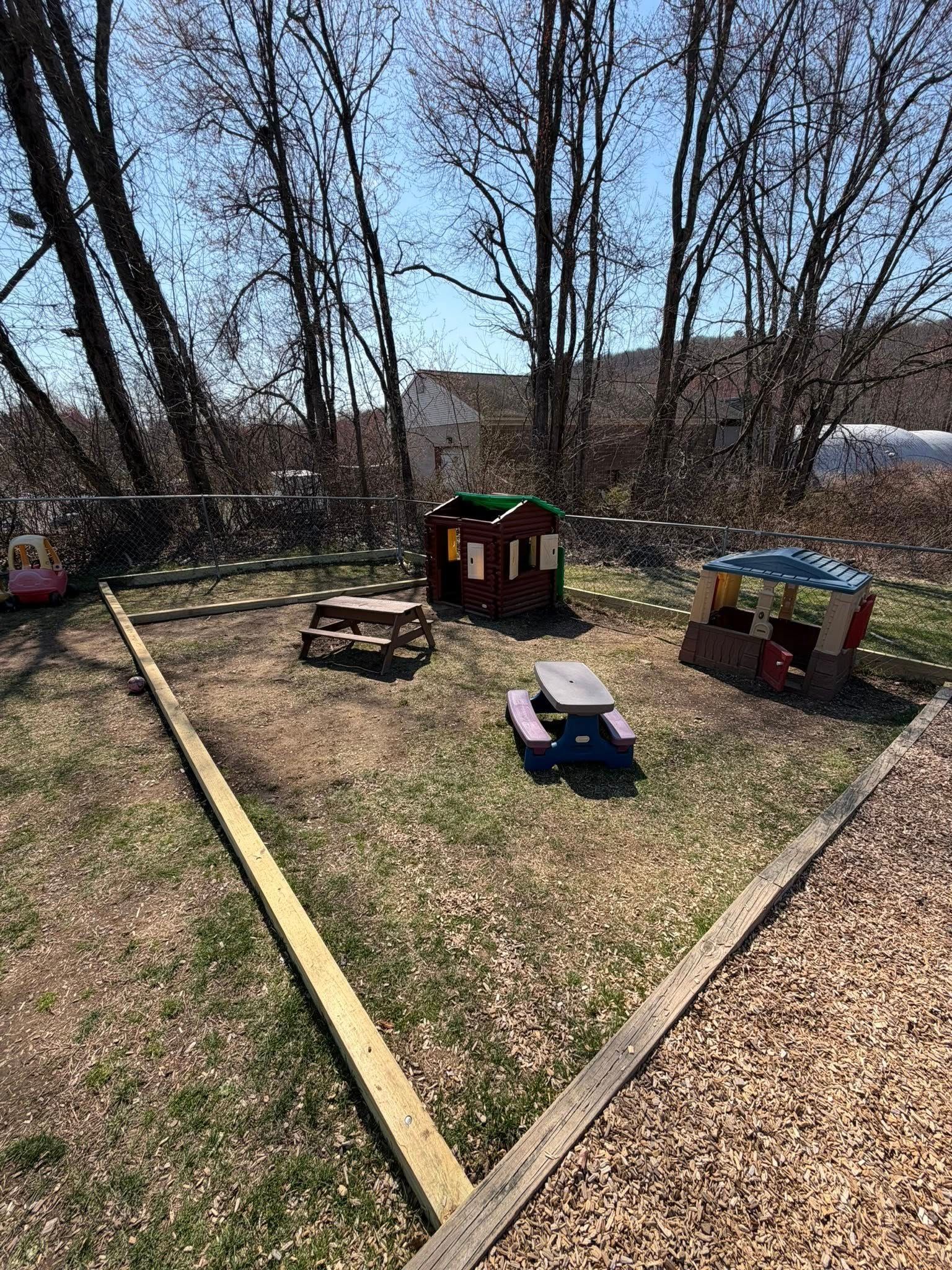 Backyard with a small play area, toy vehicles, and a wooden playhouse among leafless trees.