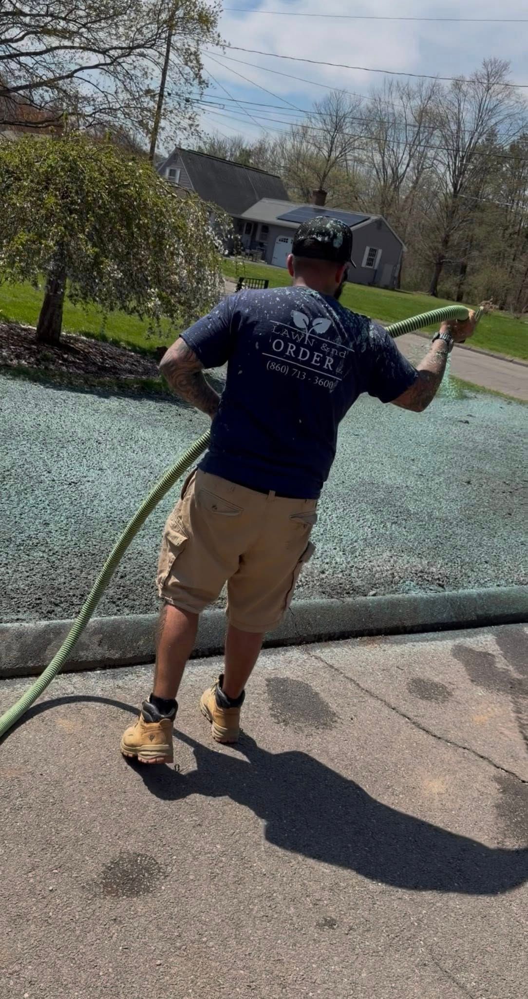 Person in a navy shirt and tan shorts sprays water from a hose across a gravel path in a sunny garden.
