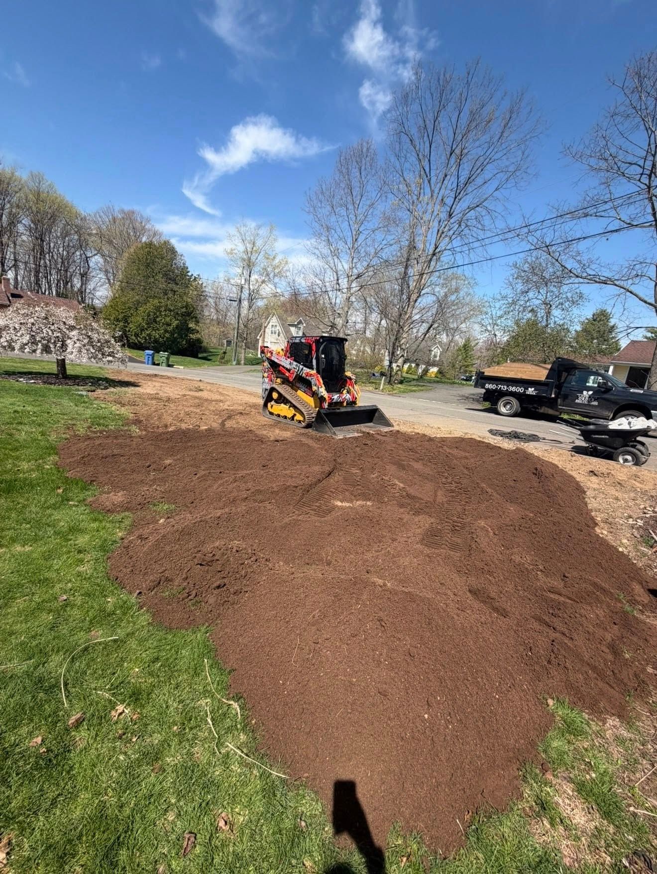 Worker on a compact loader spreading a large pile of mulch in a grassy yard on a sunny day.