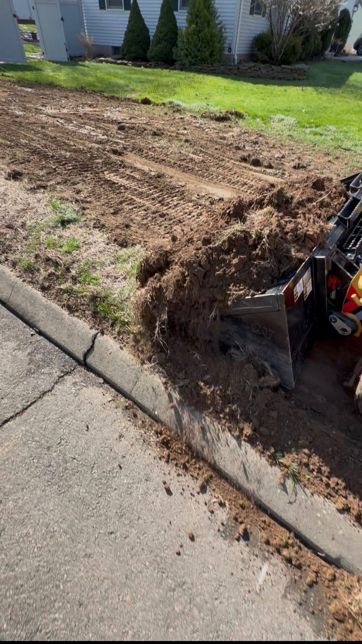 Freshly dug trench beside a sidewalk and curb with a small excavator on a lawn.