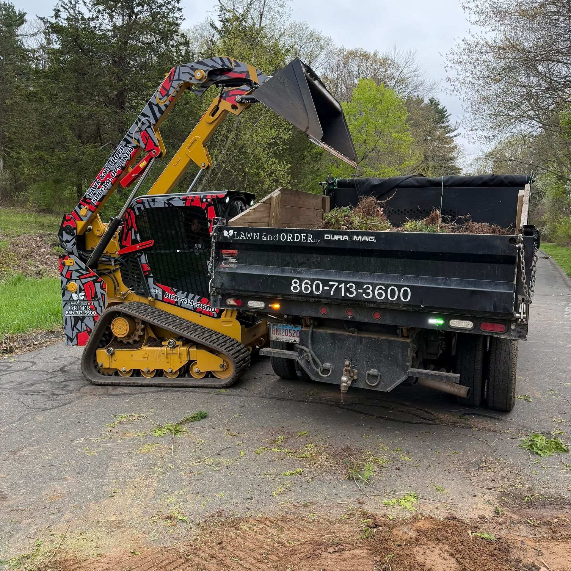 Mini excavator loading debris into a black dump truck on a gravel path, with trees in the background
