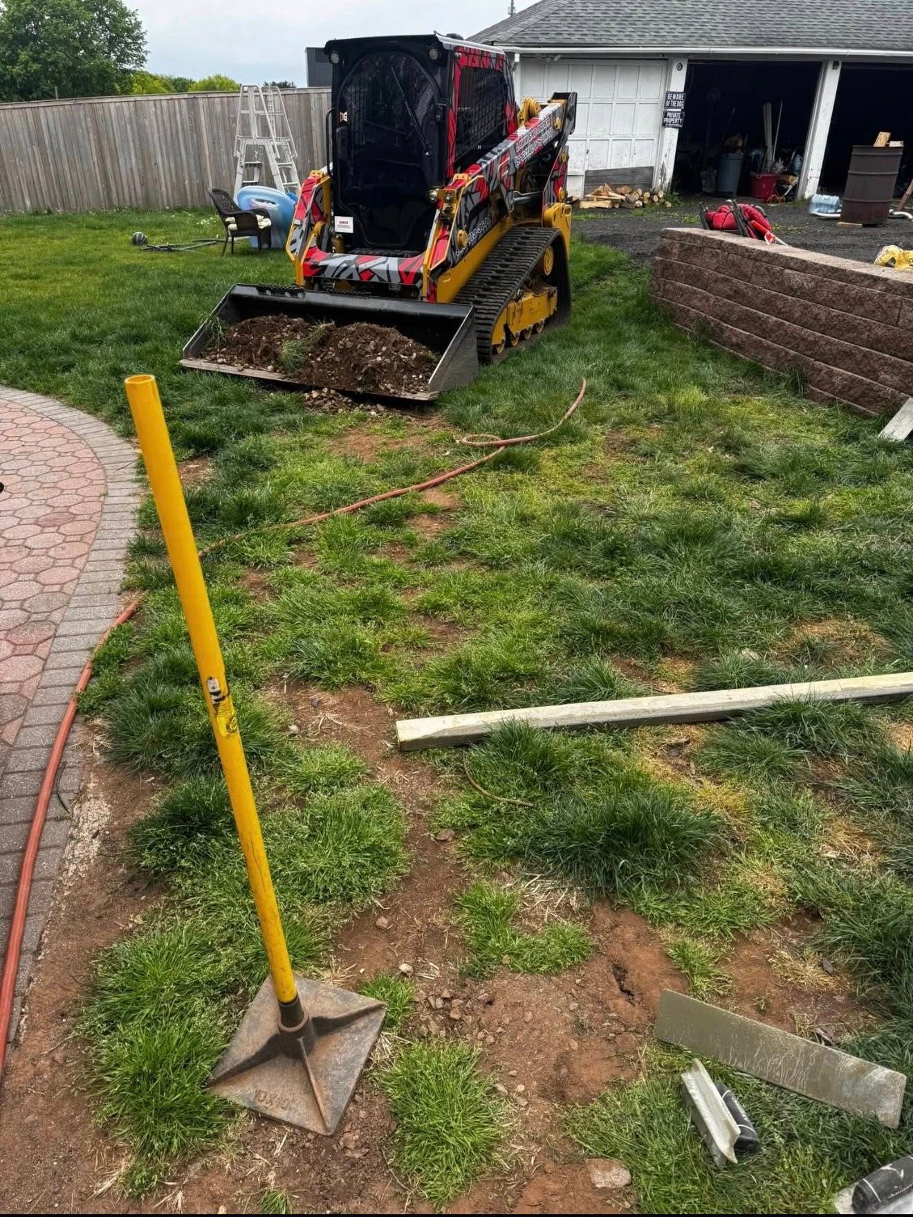 Backyard with a compact loader, yellow survey stakes, and disturbed grass near a patio and shed.