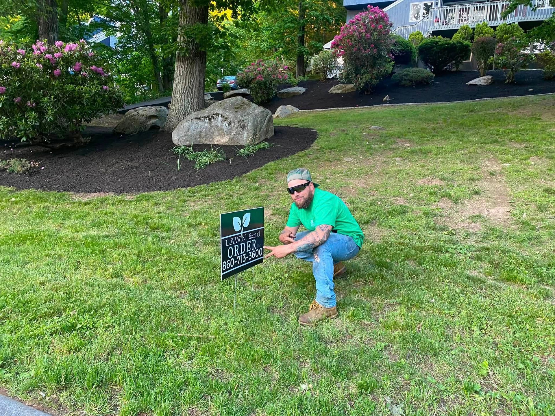 Man crouching on a lawn next to a small sign in a garden with trees and flowers.