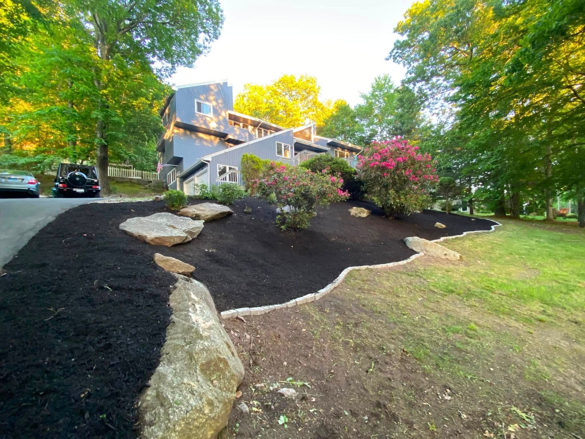 Landscaped yard with black mulch, large rocks, pink flowers, and a house in the background